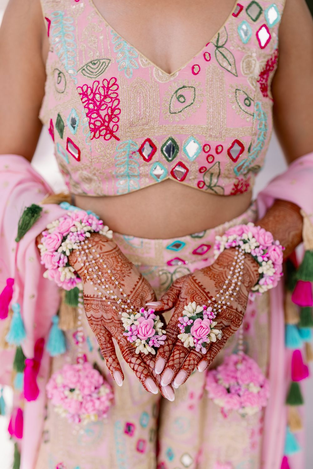 Close-up of henna decorated hands adorned with pink flowers and pearls against a pink floral background.