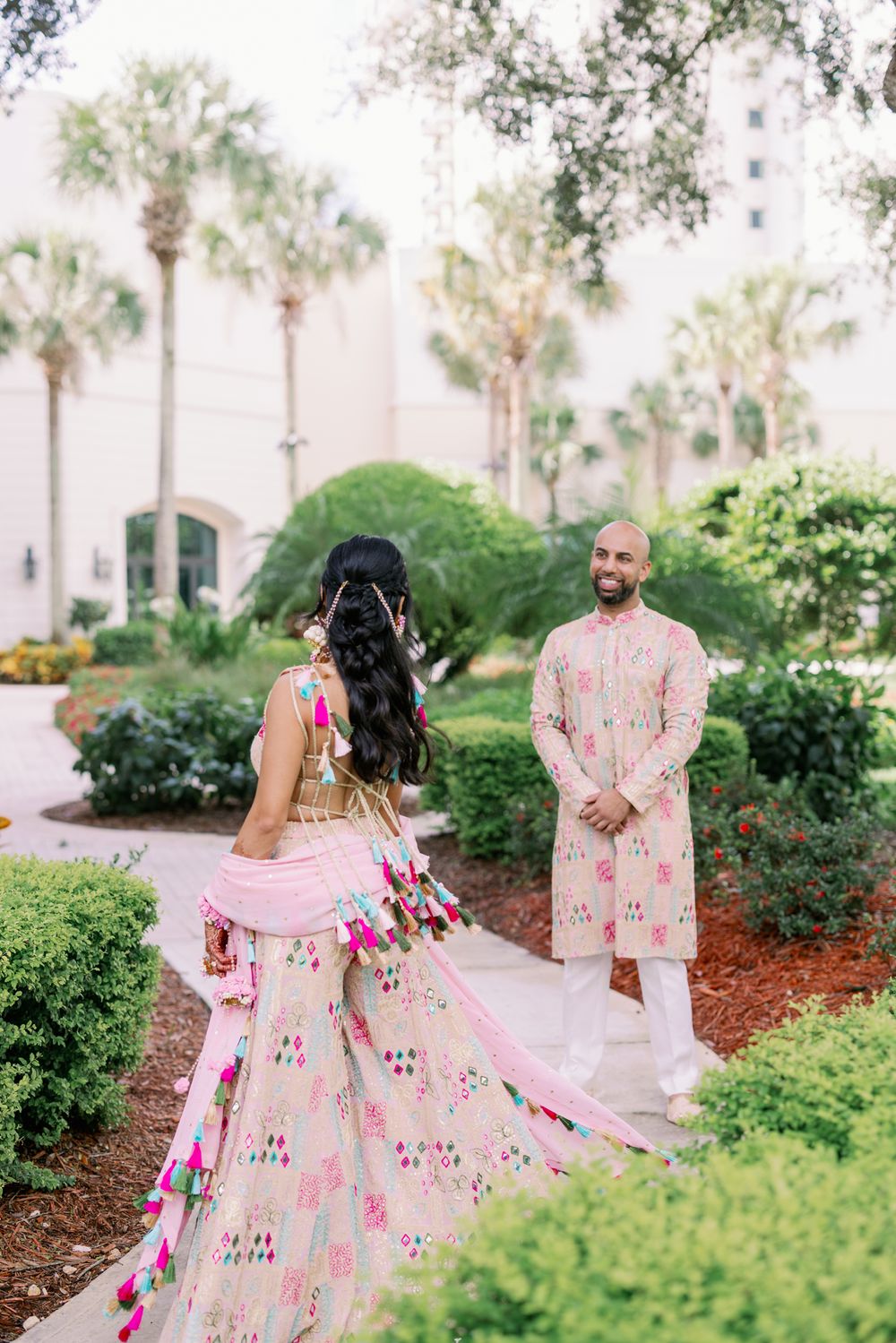 A couple in traditional Indian wedding attire poses in a lush garden setting with palm trees and greenery.