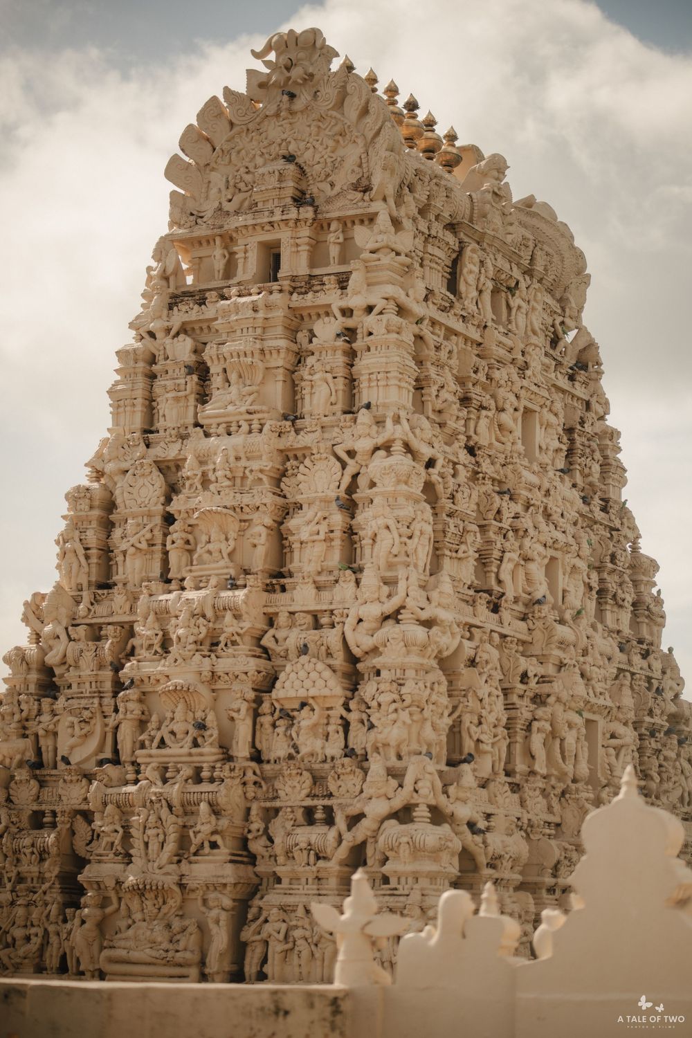 Intricate stone carvings adorn a towering South Indian Hindu temple gopuram against a cloudy sky.