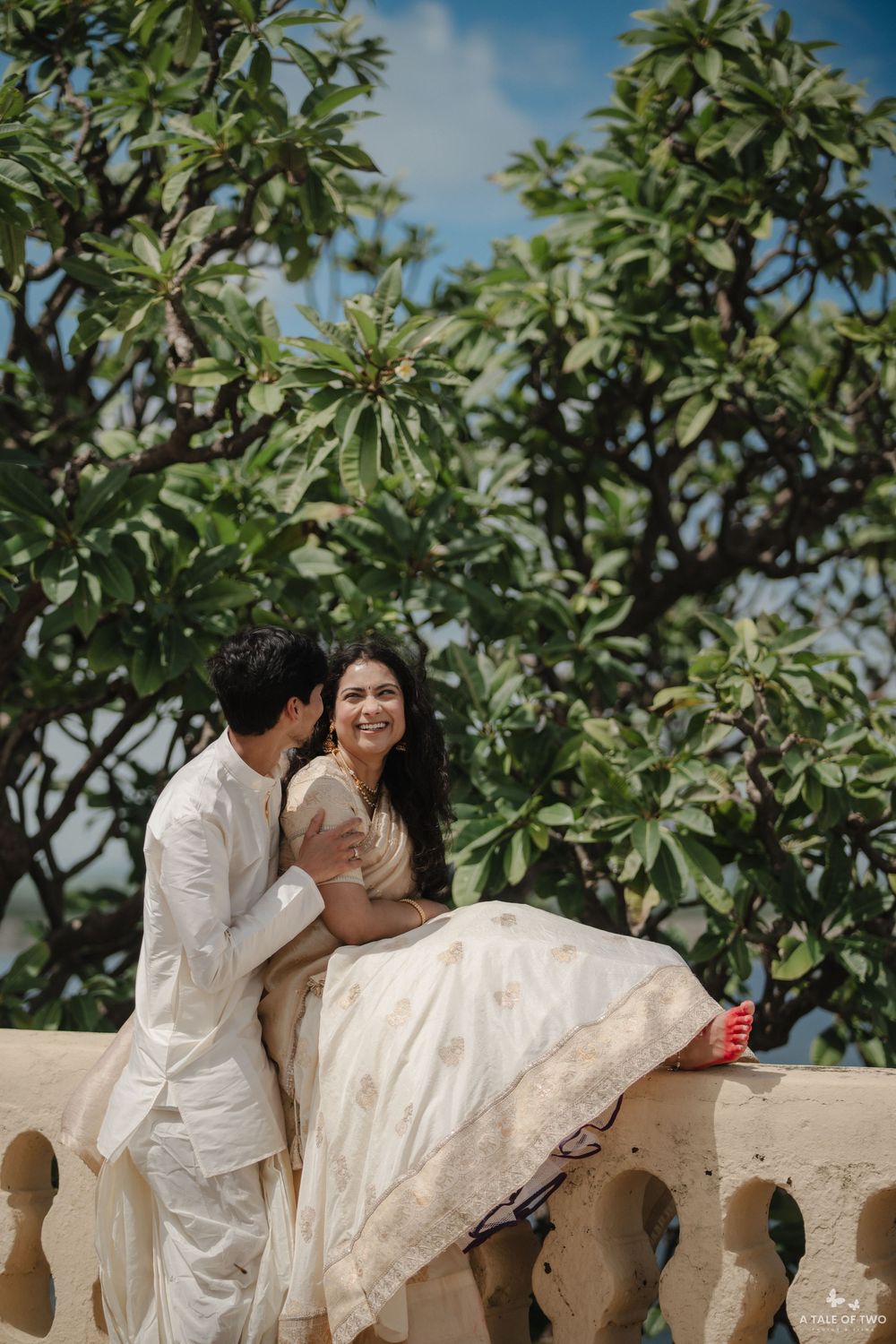 Couple in traditional Indian attire sharing a romantic moment on a terrace with lush green trees in the background.
