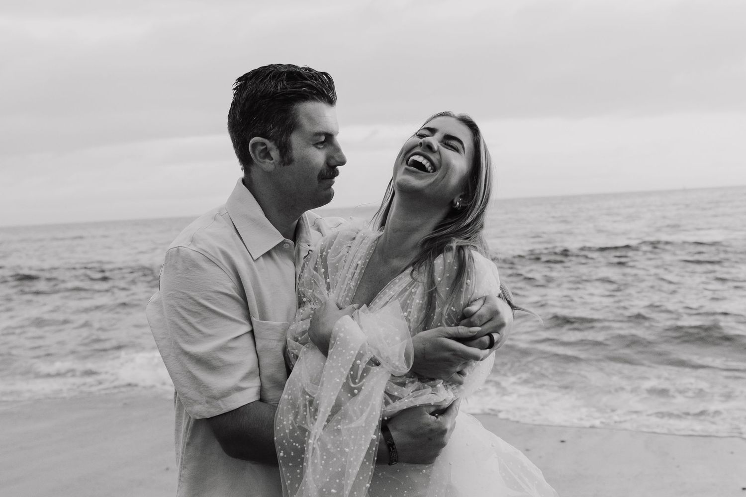 Romantic black and white photo series of a couple laughing and embracing on a beach at sunset.