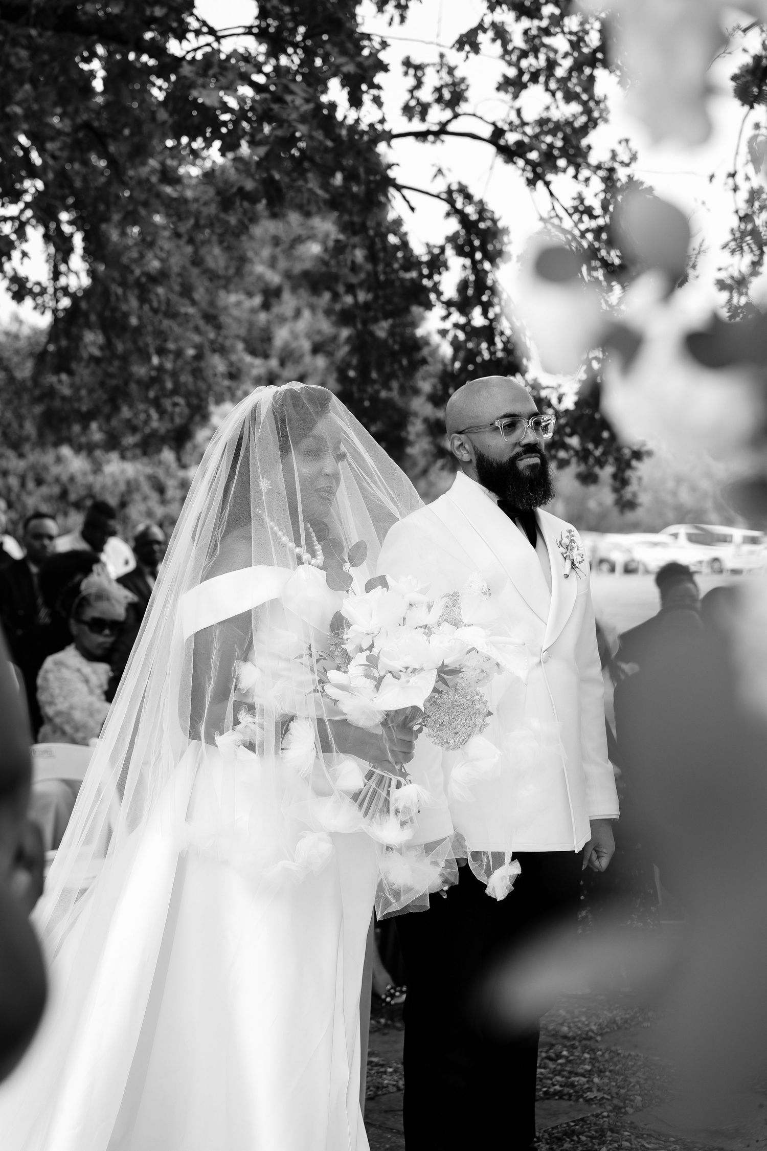 A bride in a flowing white gown walks down a cypress-lined path at dusk with illuminated garden lights.