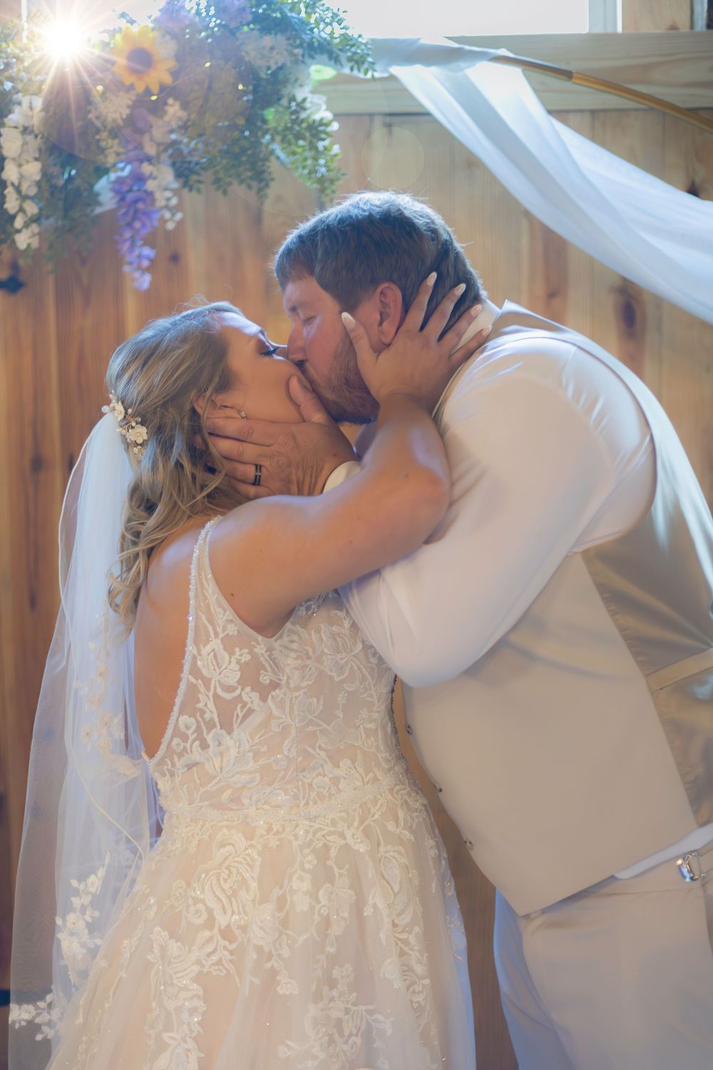 Romantic wedding kiss under floral arch with sunlight streaming through decorations.