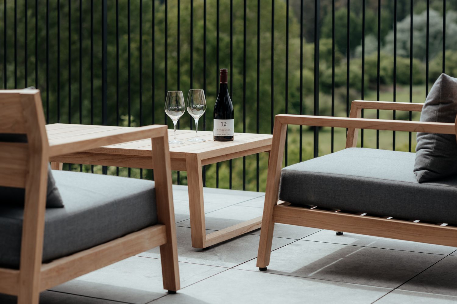 Modern outdoor wooden patio furniture with grey cushions arranged on a deck near metal railings at The Runholder by August and Vine