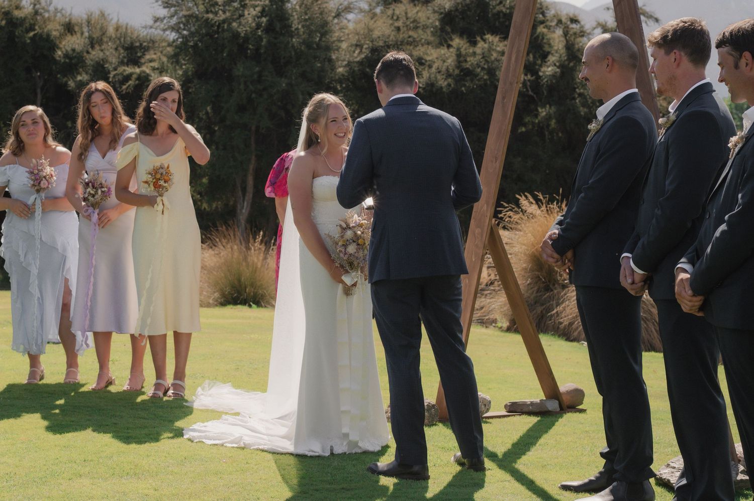 Wedding party members in formal attire stand in a line outdoors on a sunny day during a ceremony.