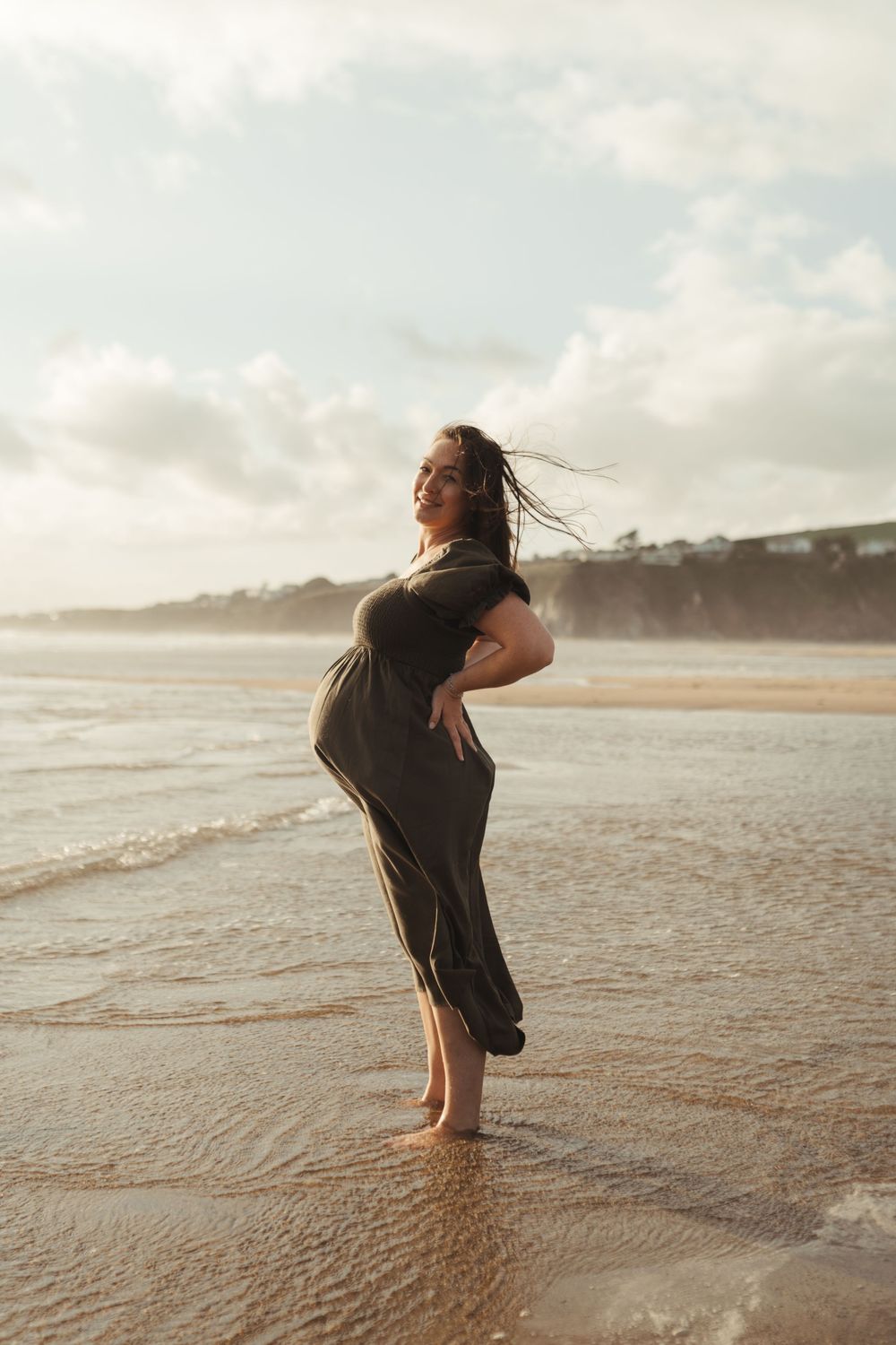 A silhouette stands gracefully on a misty beach at sunset, dress flowing in the coastal breeze.