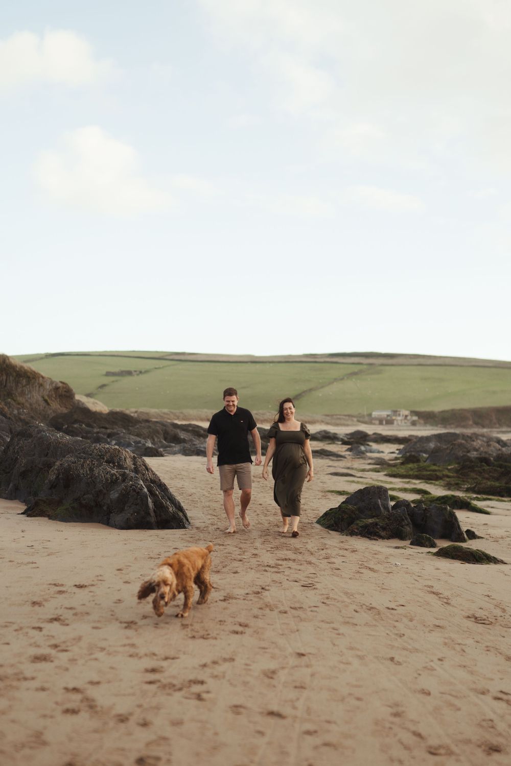 Two people walking with a golden dog on a scenic beach with rocky cliffs and green hills in the background.