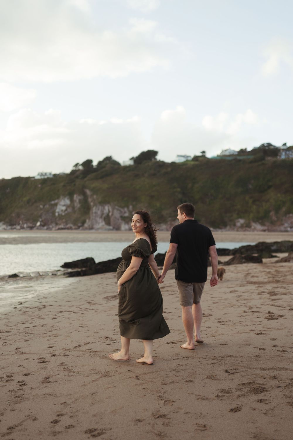 A couple walks barefoot together along a scenic beach with cliffs and rocks in the background.
