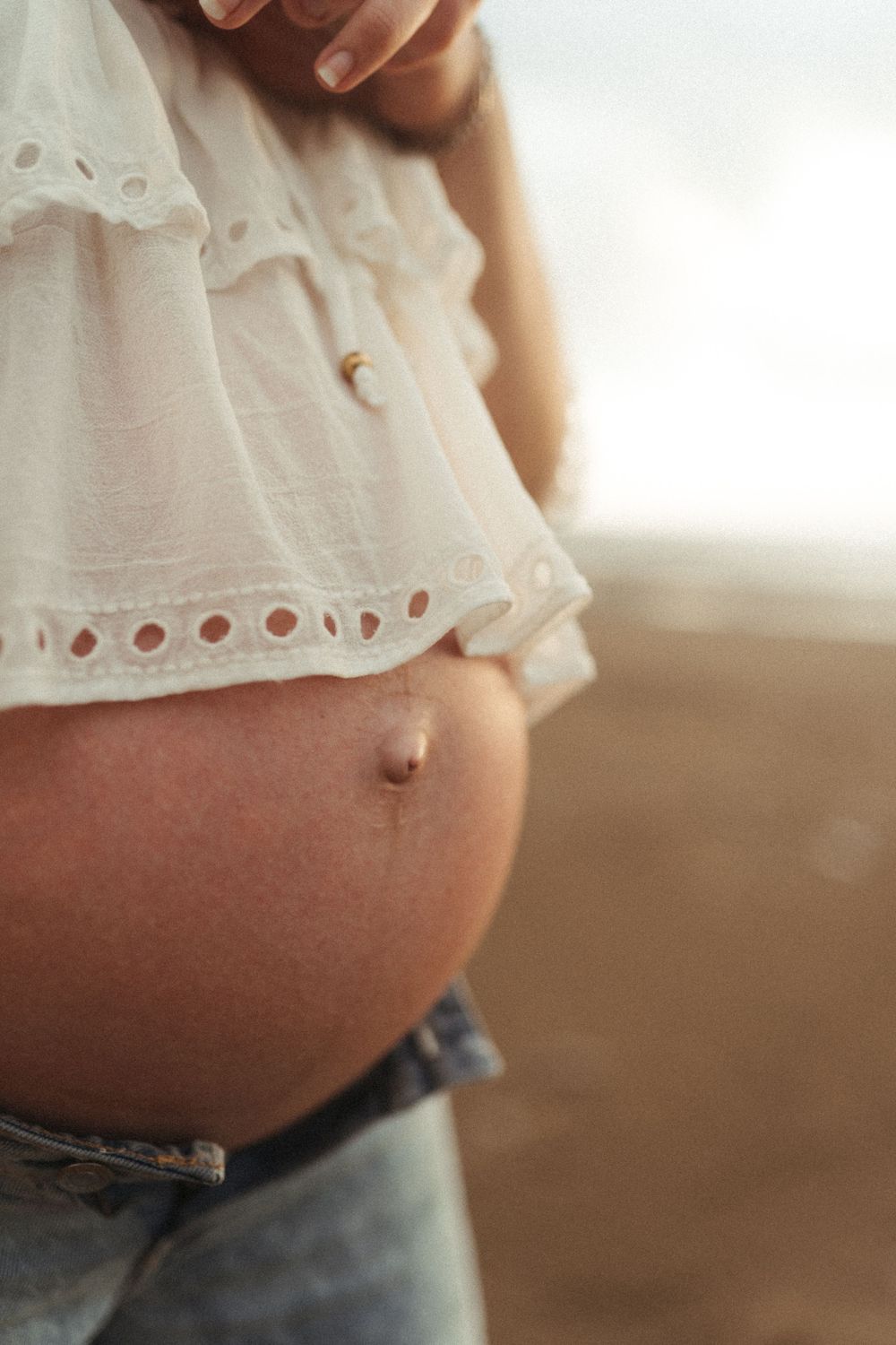 Close-up of a pregnant belly wearing a white eyelet lace top against a warm sunlit background.