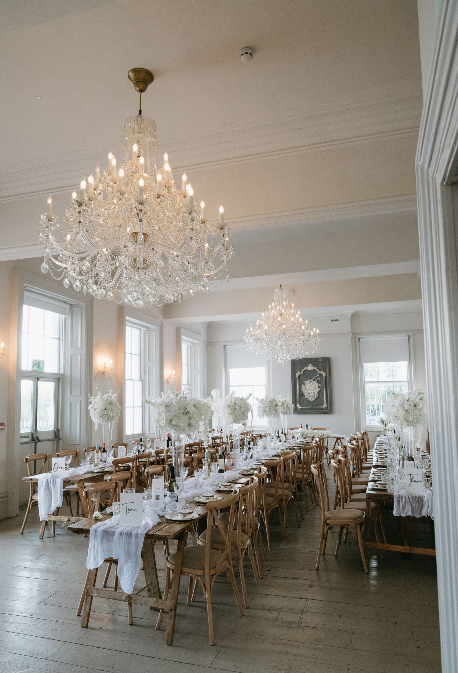 Elegant banquet hall with crystal chandeliers, wooden chairs, and white-clothed tables arranged in neat rows.
