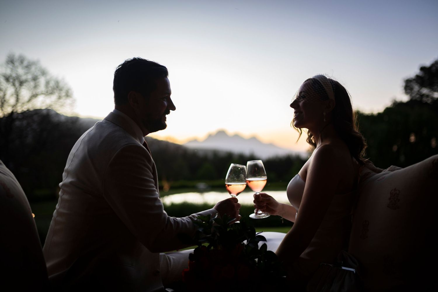 Two silhouettes share wine glasses at sunset, creating a romantic outdoor moment against a darkening sky.