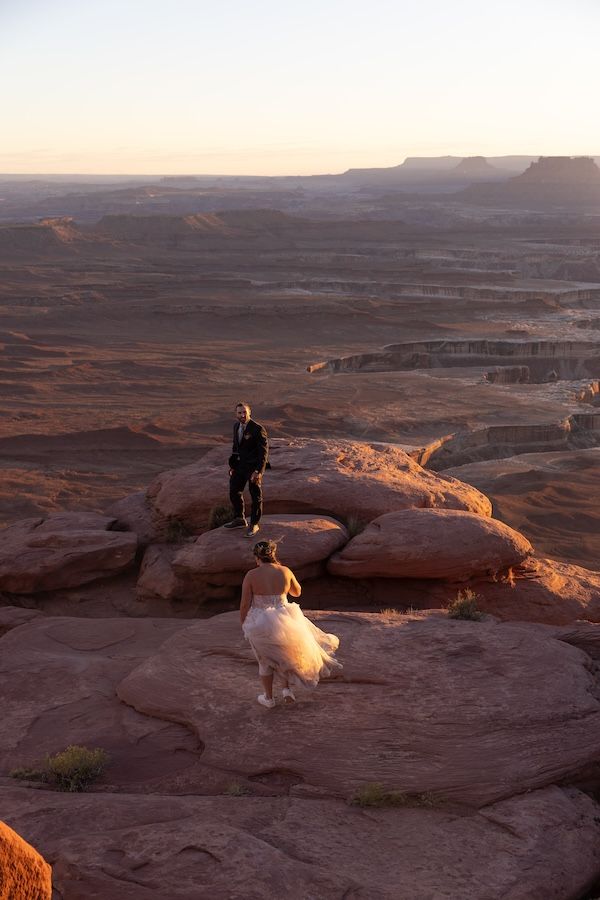 A person stands on red rock formations overlooking a vast desert canyon landscape at sunset in Utah.