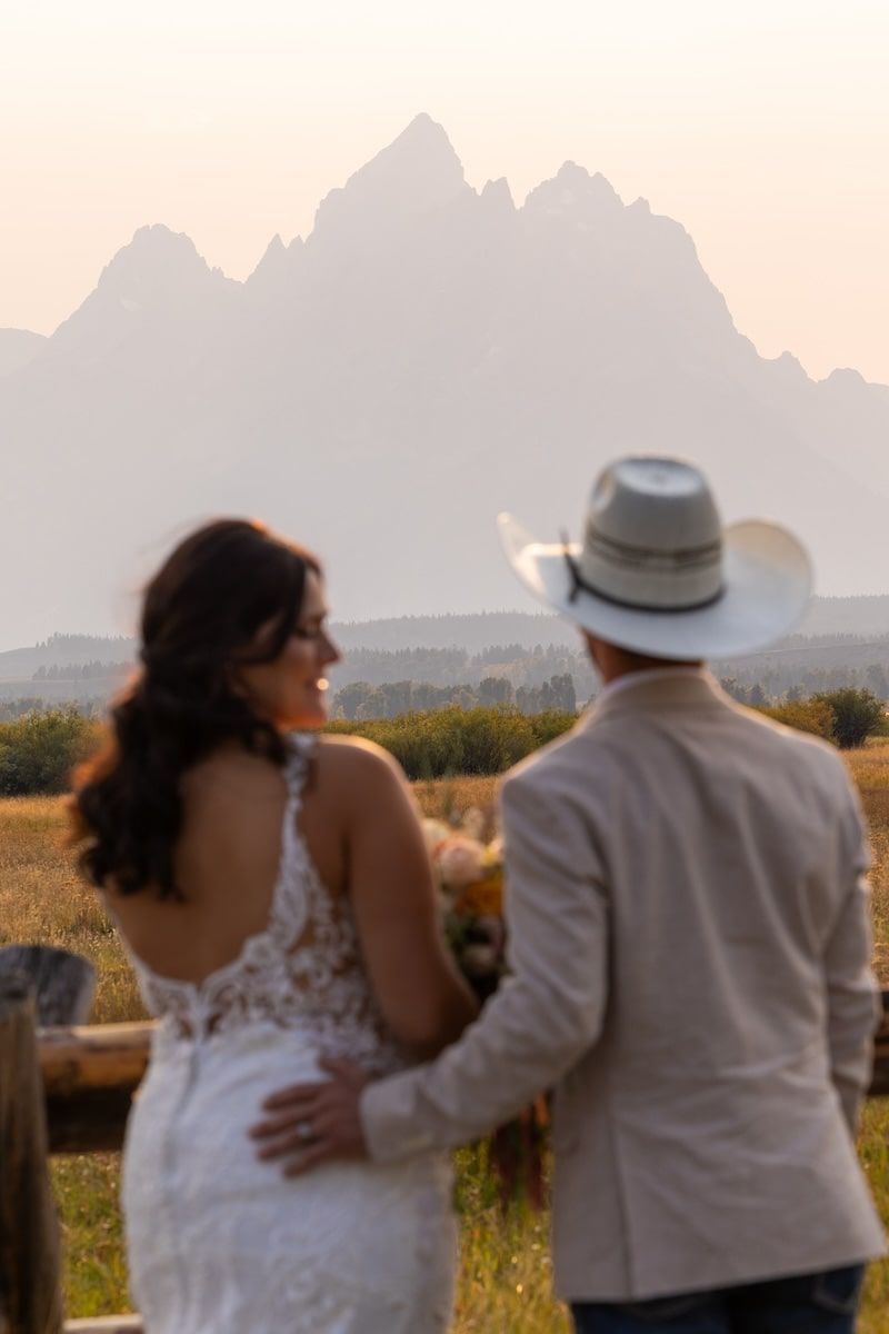 A couple in white clothing embraces while watching the sunset over mountain peaks and wildflower fields.
