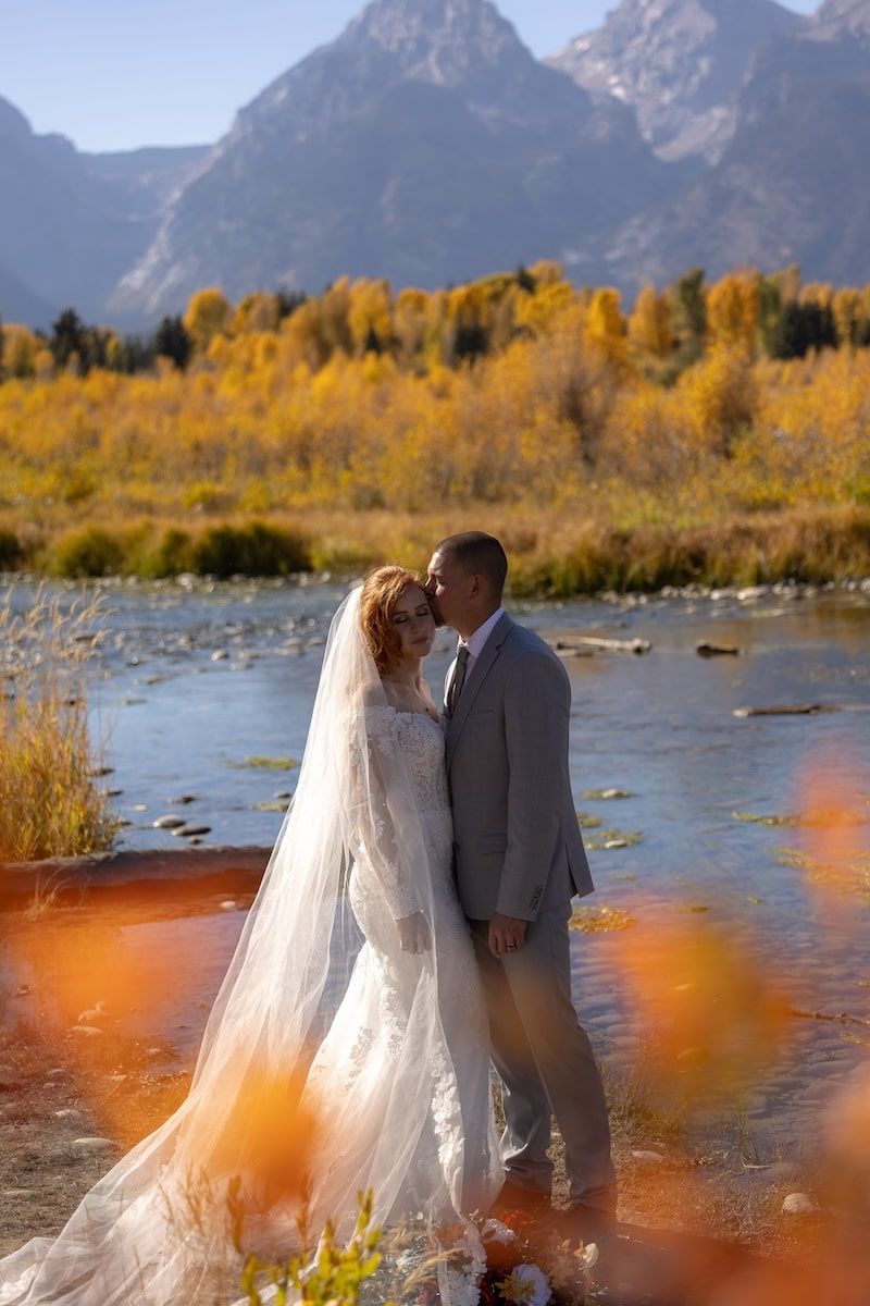 Couple in wedding attire embracing by a river with golden autumn trees and mountain peaks in Grand Teton National Park.
