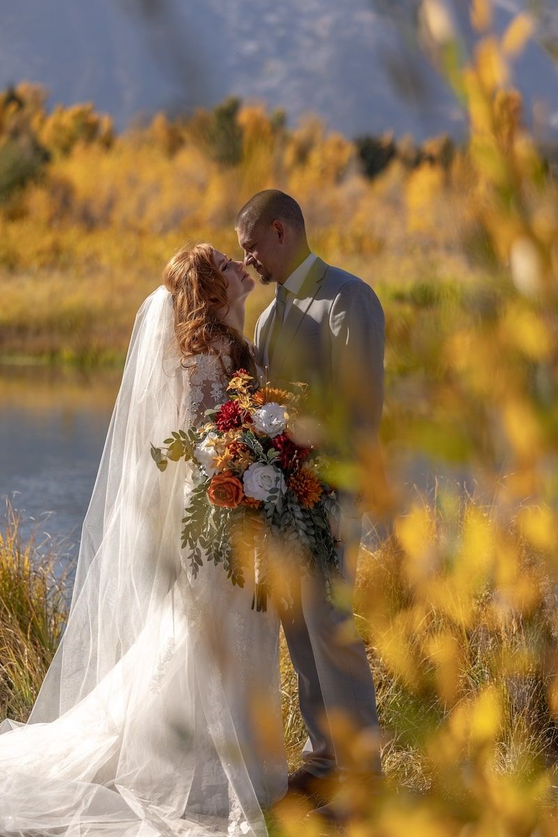 Romantic autumn wedding couple embraces by lakeside during golden hour with flowing white veil and fall bouquet.