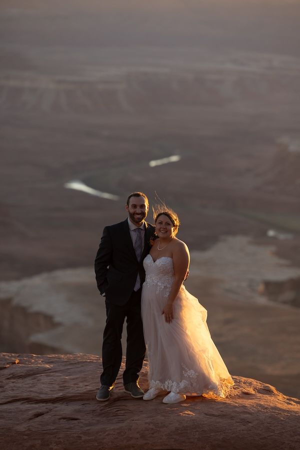 A couple in wedding attire poses at sunset on a rocky desert overlook.