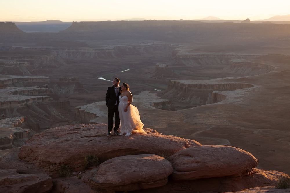 Panoramic view of a desert canyon landscape at sunset with dramatic lighting and silhouetted figures on the cliffs.
