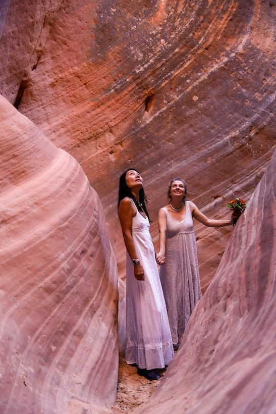 Two people in white dresses pose against curved red sandstone canyon walls in dramatic lighting.