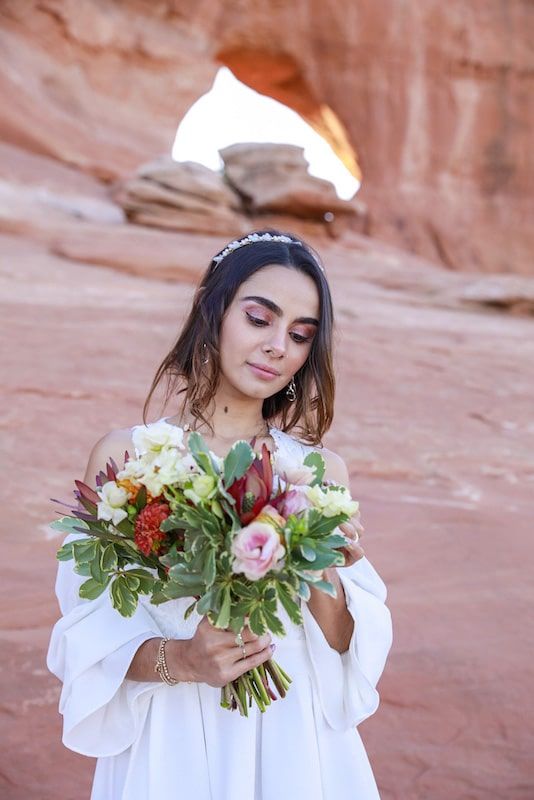 A bride in a white dress holds a colorful bouquet with pink and red flowers against red rock canyon walls.