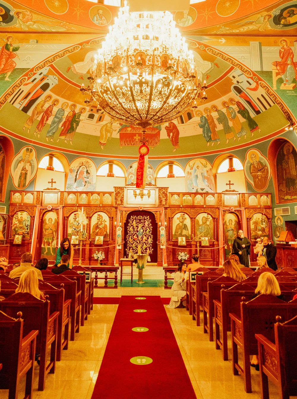 Panoramic view of an ornate Orthodox church interior with crystal chandeliers, red carpet, and wooden pews.