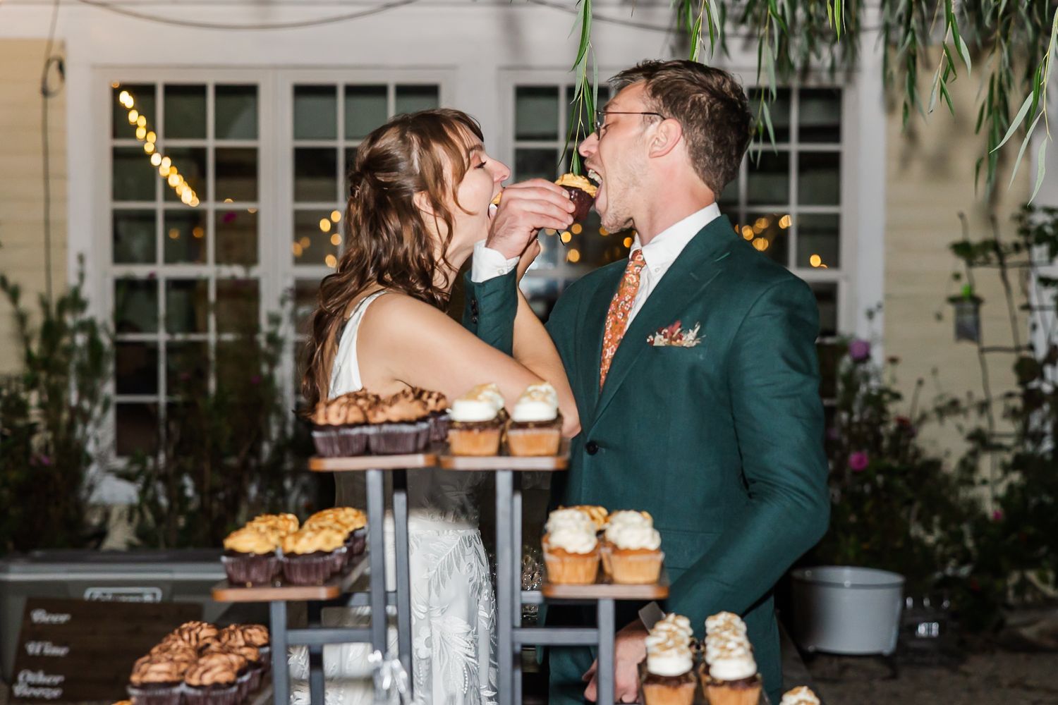 A couple shares an intimate moment at a rustic wedding reception dessert table with cupcakes and treats.