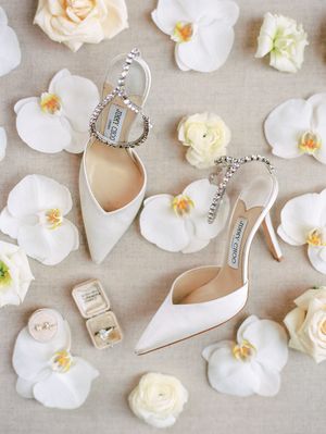 White orchids and roses scattered among elegant bridal shoes and jewelry on a neutral linen background.