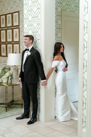 A couple in wedding attire peek around opposite sides of a white column in a playful first look sequence.