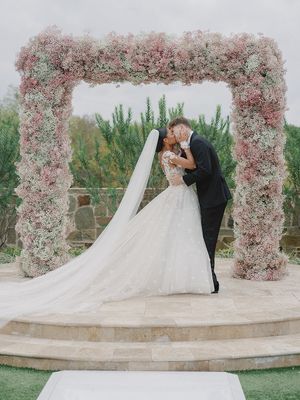 Romantic wedding couple embraces under a stunning pink floral arch during an outdoor ceremony.