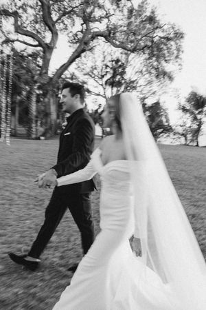 A black and white wedding photo of a couple walking outdoors, with the bride wearing a flowing white veil and gown.