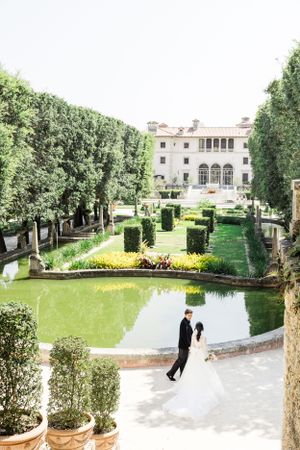 Elegant couple walking through manicured garden with mansion and reflecting pool.