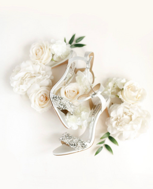 Elegant bridal shoes surrounded by soft white roses and delicate foliage on white background.