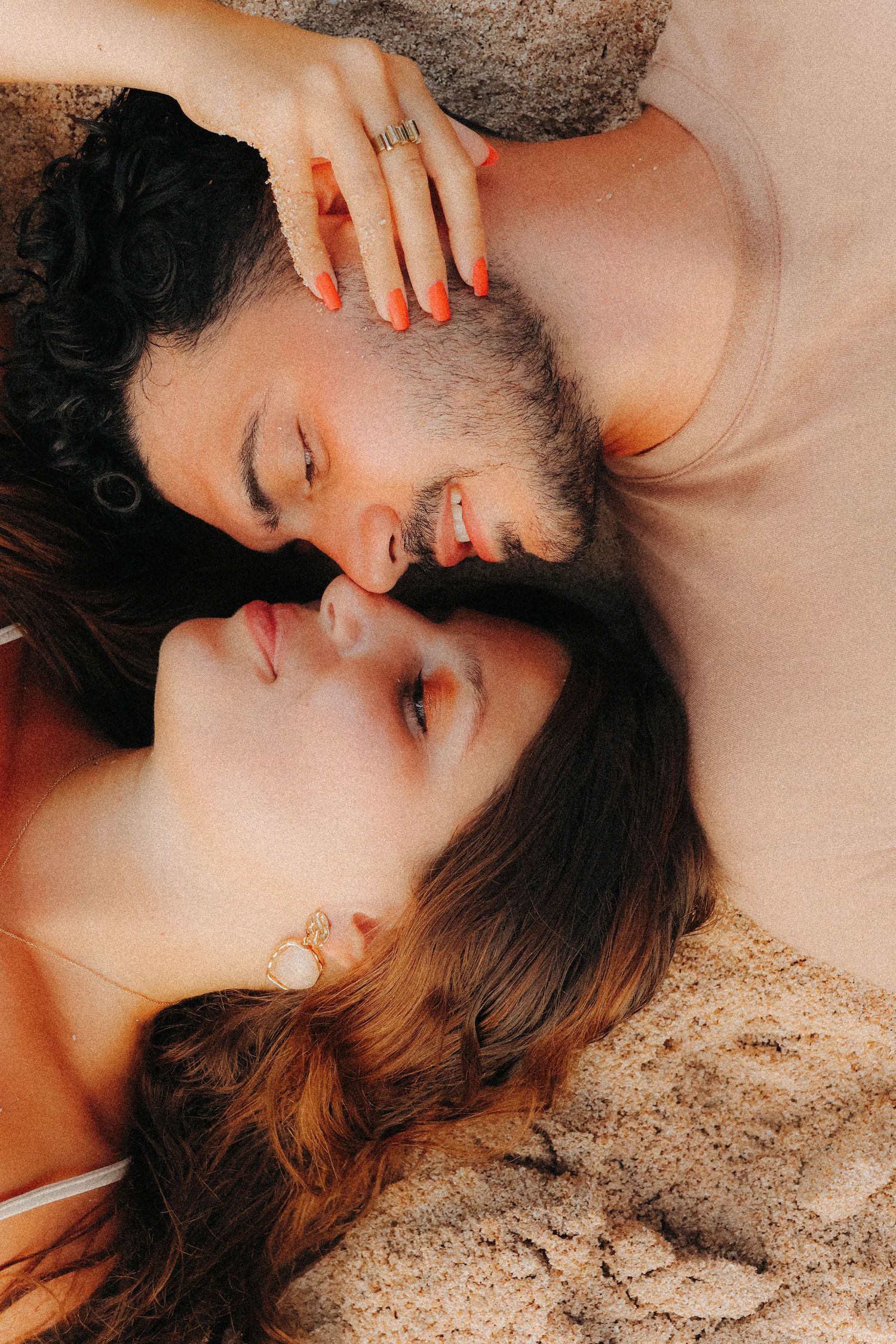 Overhead view of couple lying on sand face to face, woman with orange nails smiling softly.
