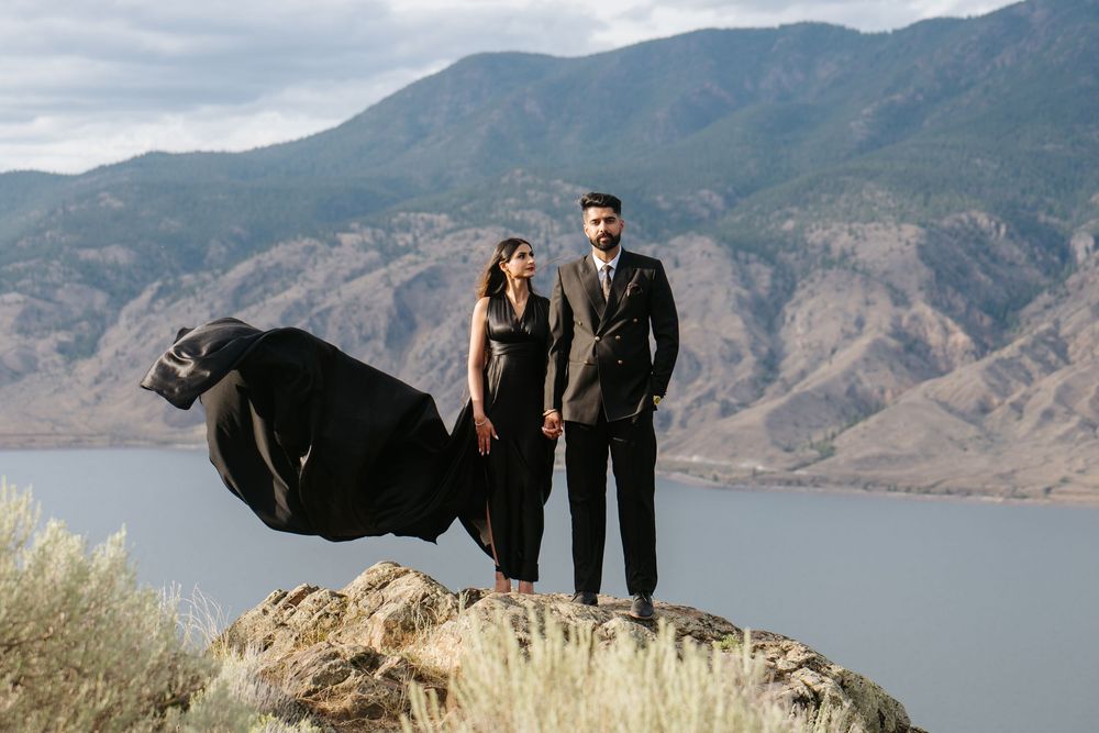 A couple poses on a scenic mountain overlook with a lake and dramatic cloudy sky in the background.