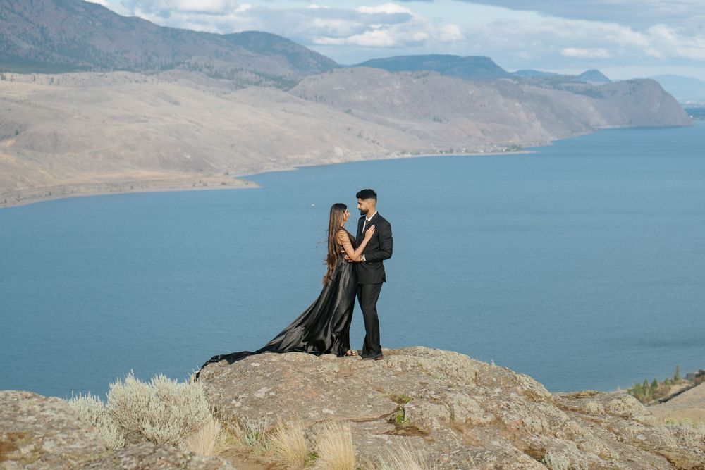 A couple in formal attire stands on a rocky cliff overlooking a blue mountain lake on a cloudy day.