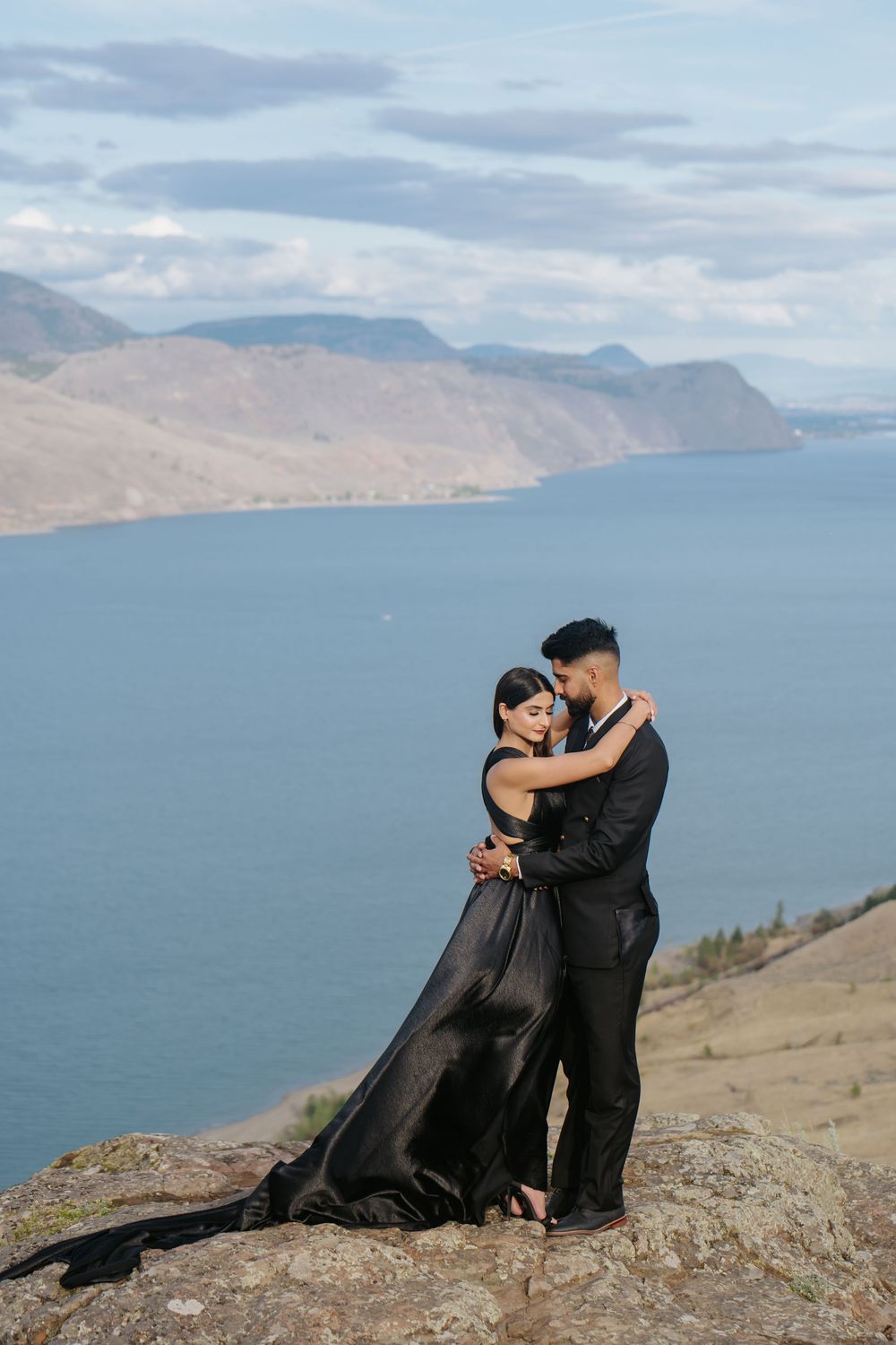 A couple embraces on a rocky cliff overlooking a scenic mountain lake landscape with cloudy skies.