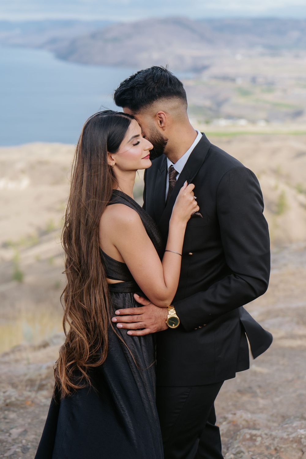 A couple shares intimate moments against a scenic mountain backdrop wearing black formal attire.