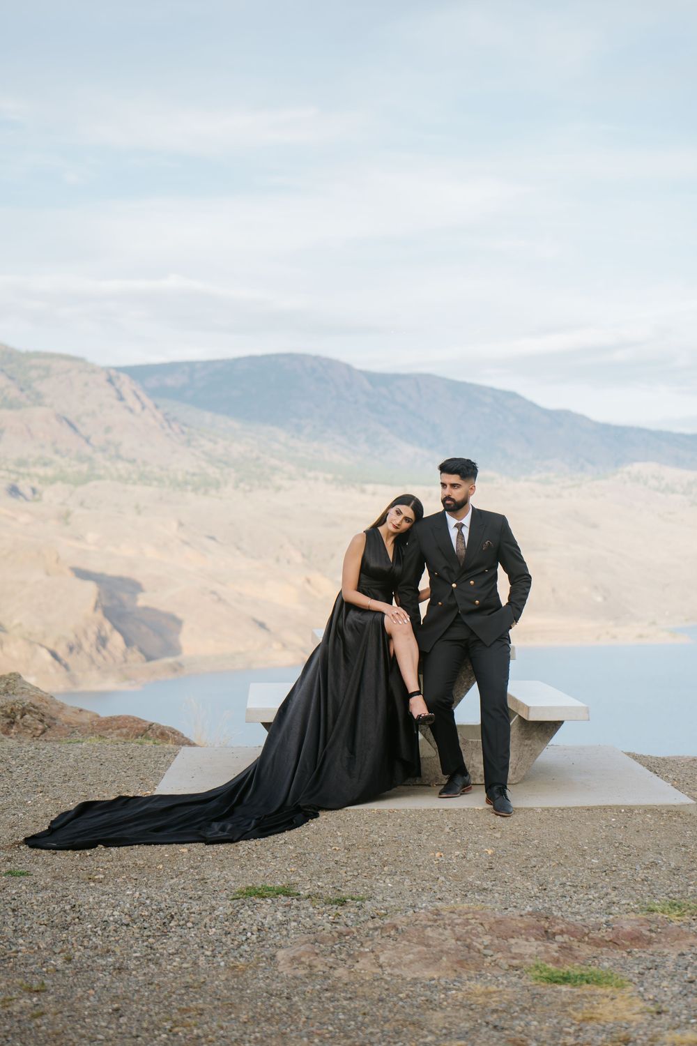A couple in formal attire poses dramatically on a rocky cliff overlooking a scenic mountain lake landscape.