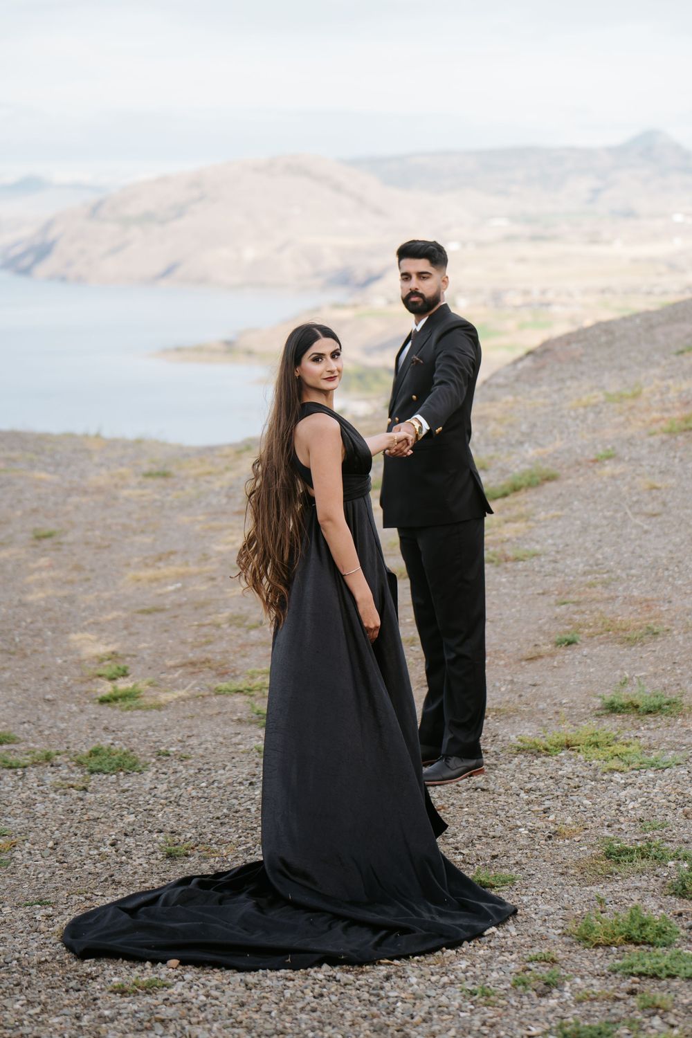 A couple in black formal wear poses dramatically on a mountainside overlooking a scenic landscape.