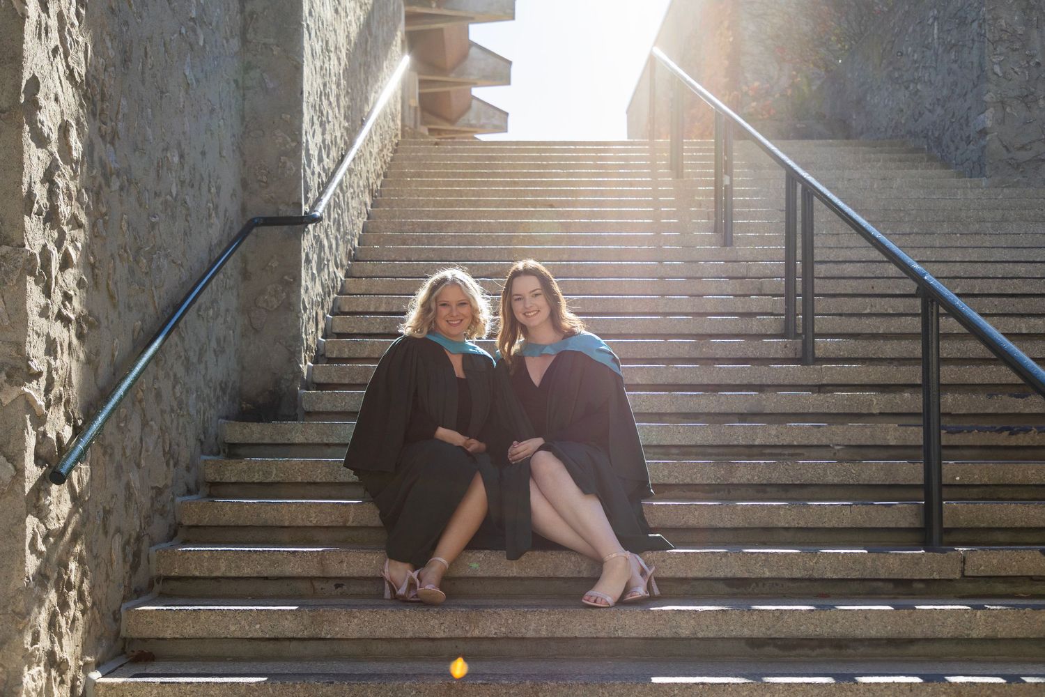 2 Female graduates of Trent University sitting on the campus stairs wearing their gowns