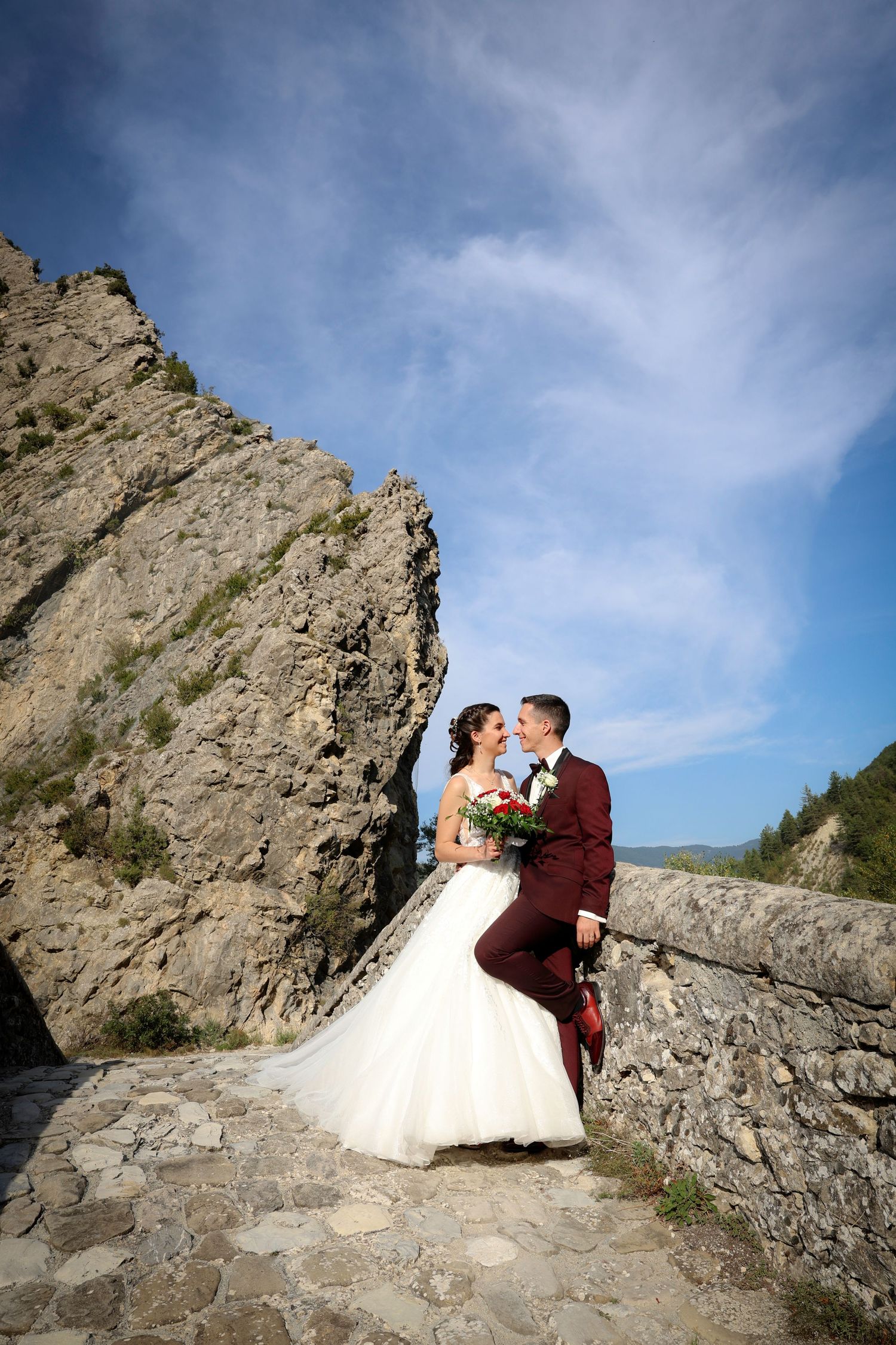 Couple in wedding attire posing on rocky cliffs against a blue sky backdrop.