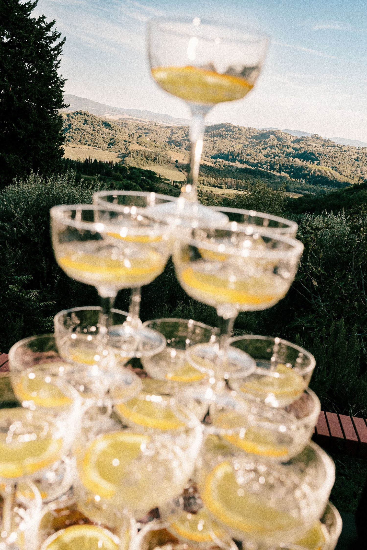 A tower of champagne coupe glasses filled with golden cocktails is set against a scenic mountain landscape backdrop.
