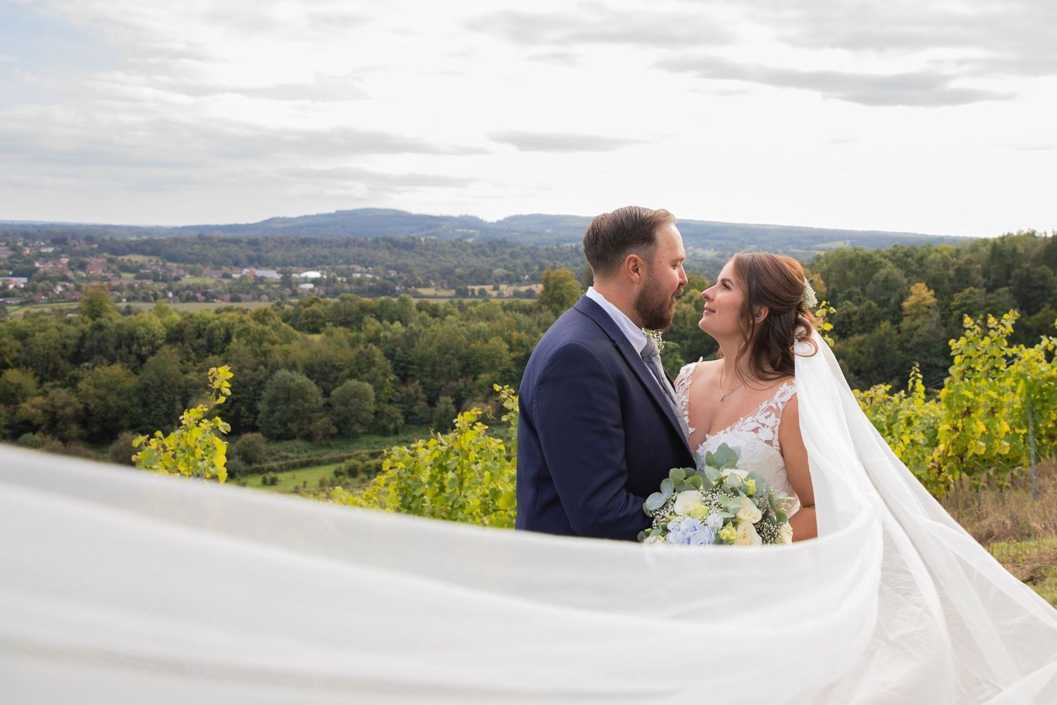 A couple shares a romantic moment overlooking a scenic hillside landscape with flowing white veil in the breeze.