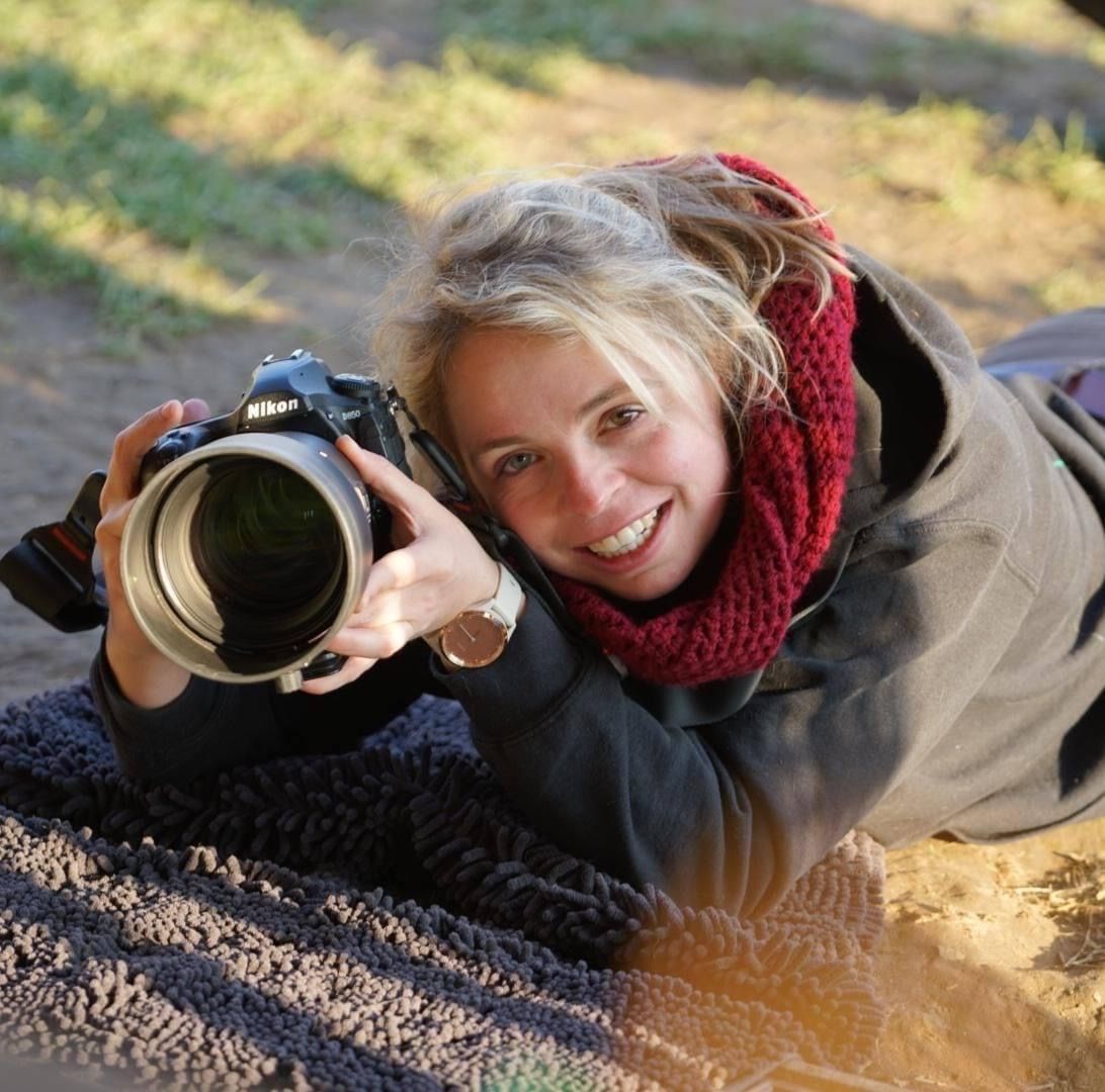 Photographer with professional camera and red scarf lying on ground while capturing outdoor shots during golden hour.