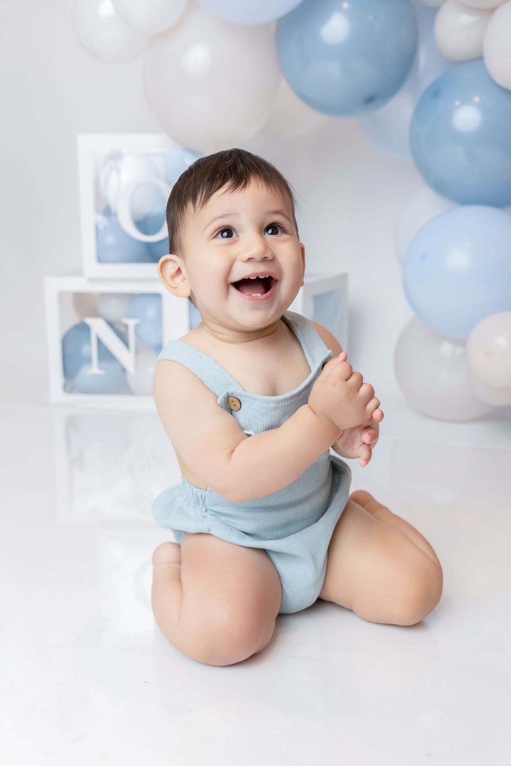 A happy baby boy in a blue romper sits smiling surrounded by blue and white balloons at a first birthday photoshoot.