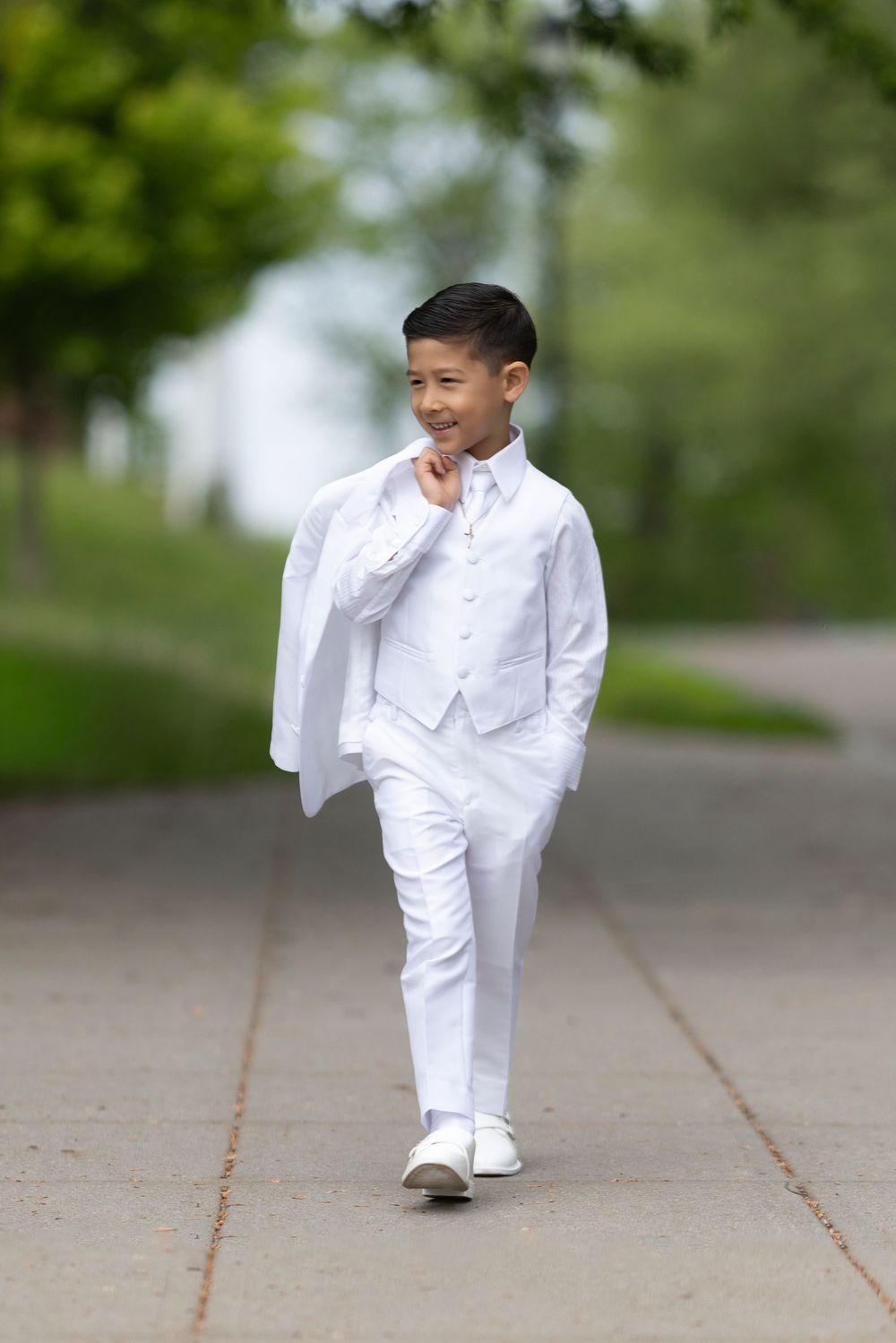 A young boy in a white suit walks confidently down a path, carrying his jacket over his shoulder.