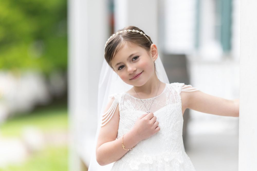 Young girl in white lace First Communion dress and veil, smiling outdoors near white pillars.