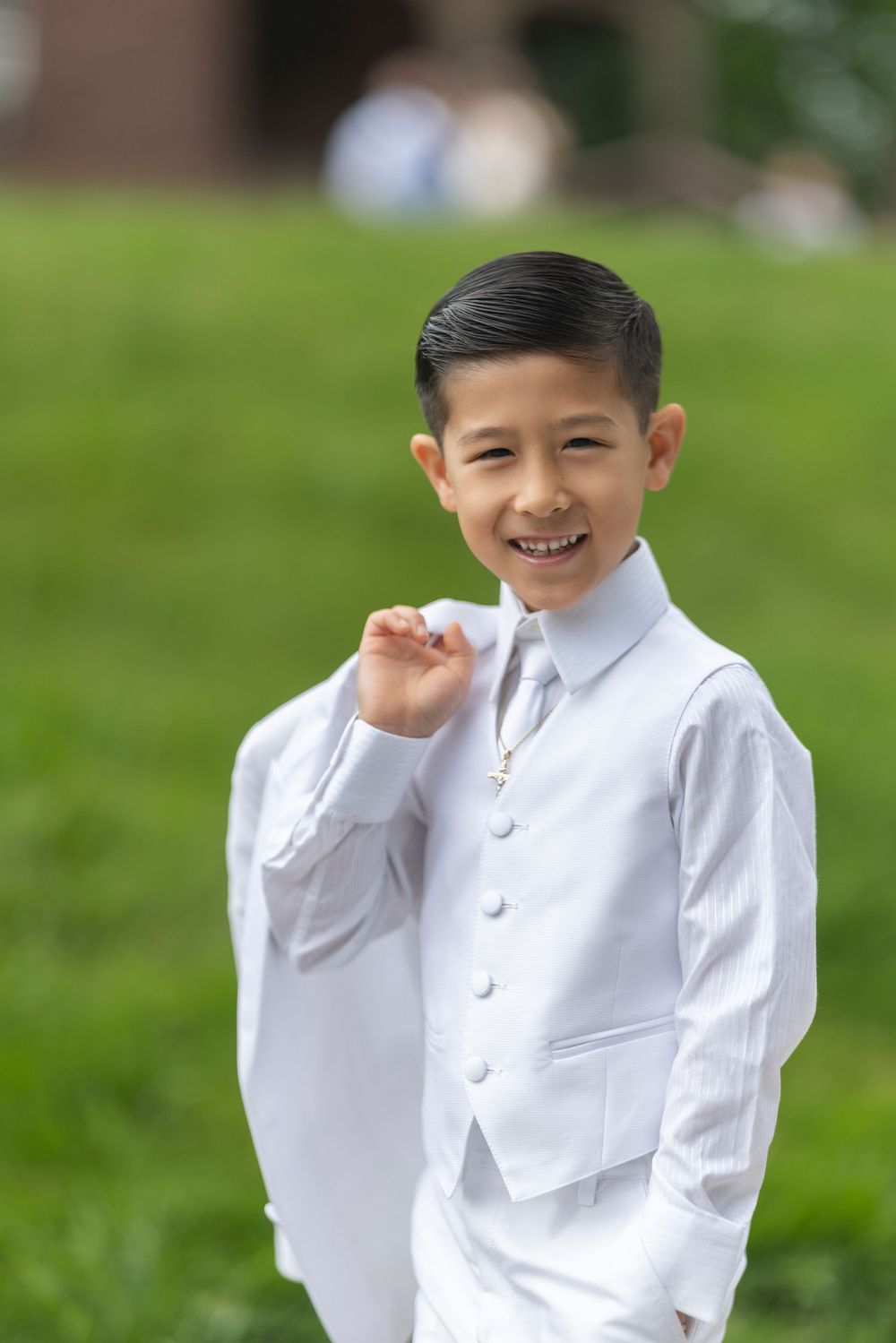 Smiling young boy in white suit and tie posing outdoors on green grass.