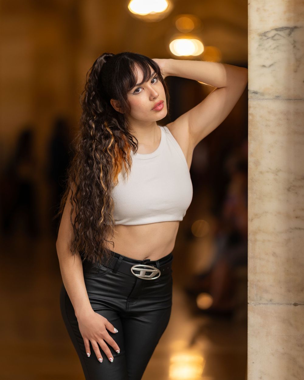Young woman in white crop top and black leather pants poses confidently by a marble column in elegant interior.