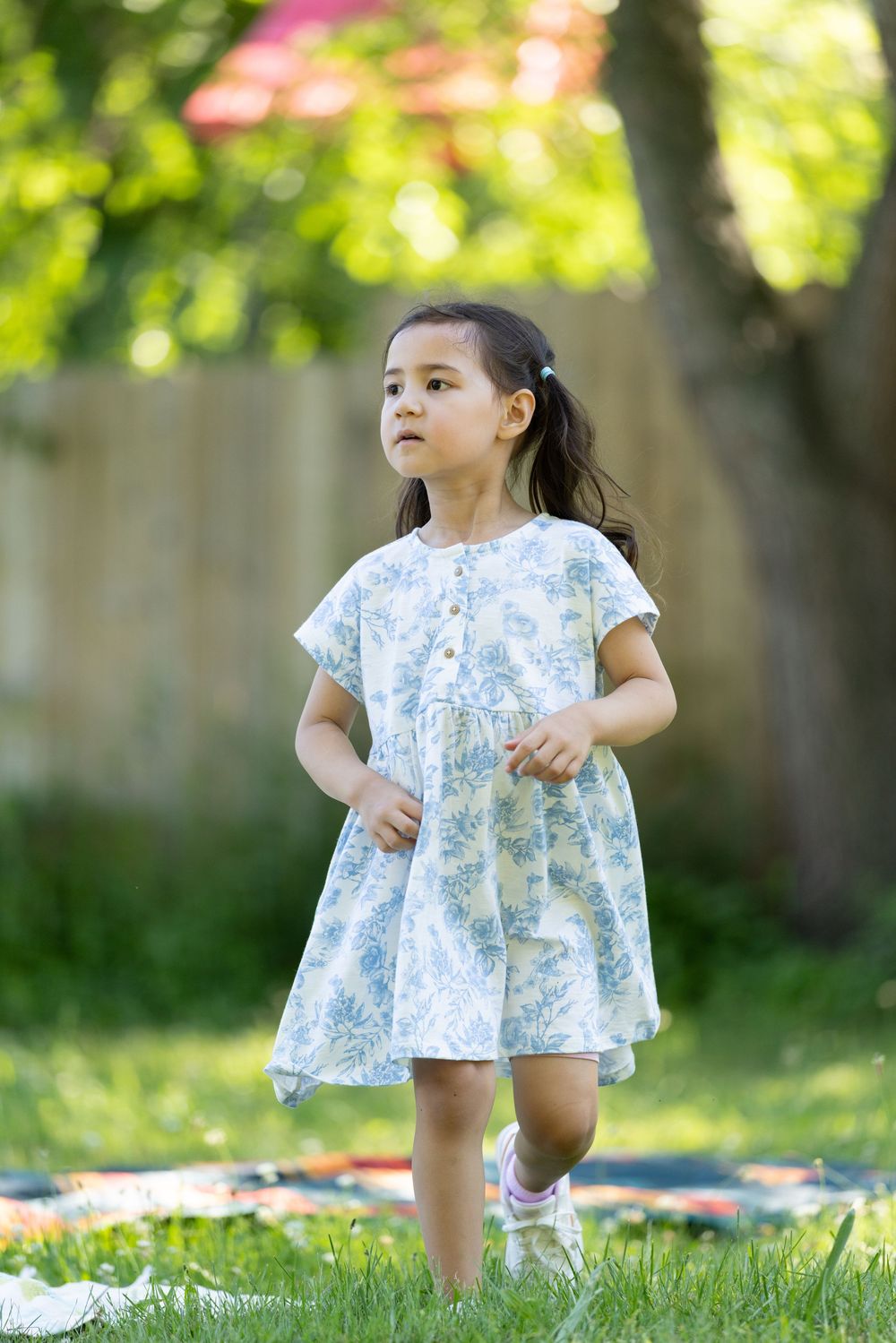 Young girl in floral dress playing in sunny backyard garden, running on green grass near wooden fence.