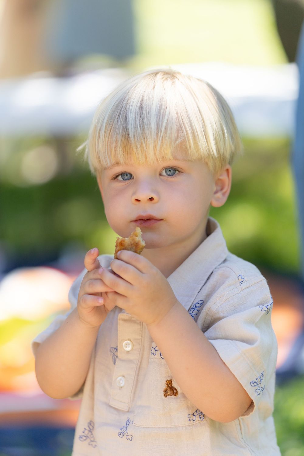 Blonde toddler boy eating a snack outdoors, wearing a light blue shirt in a sunny garden setting.